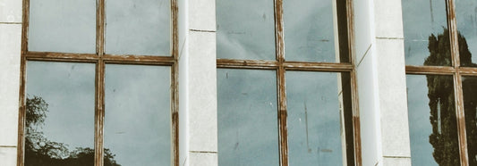Close-up of tall antique wavy windows with weathered wooden frames reflecting trees and the sky.